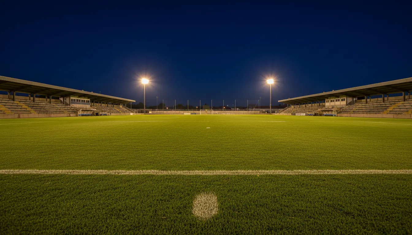 Campo de fútbol de Segunda División con césped natural y gradas modestas iluminadas por focos nocturnos